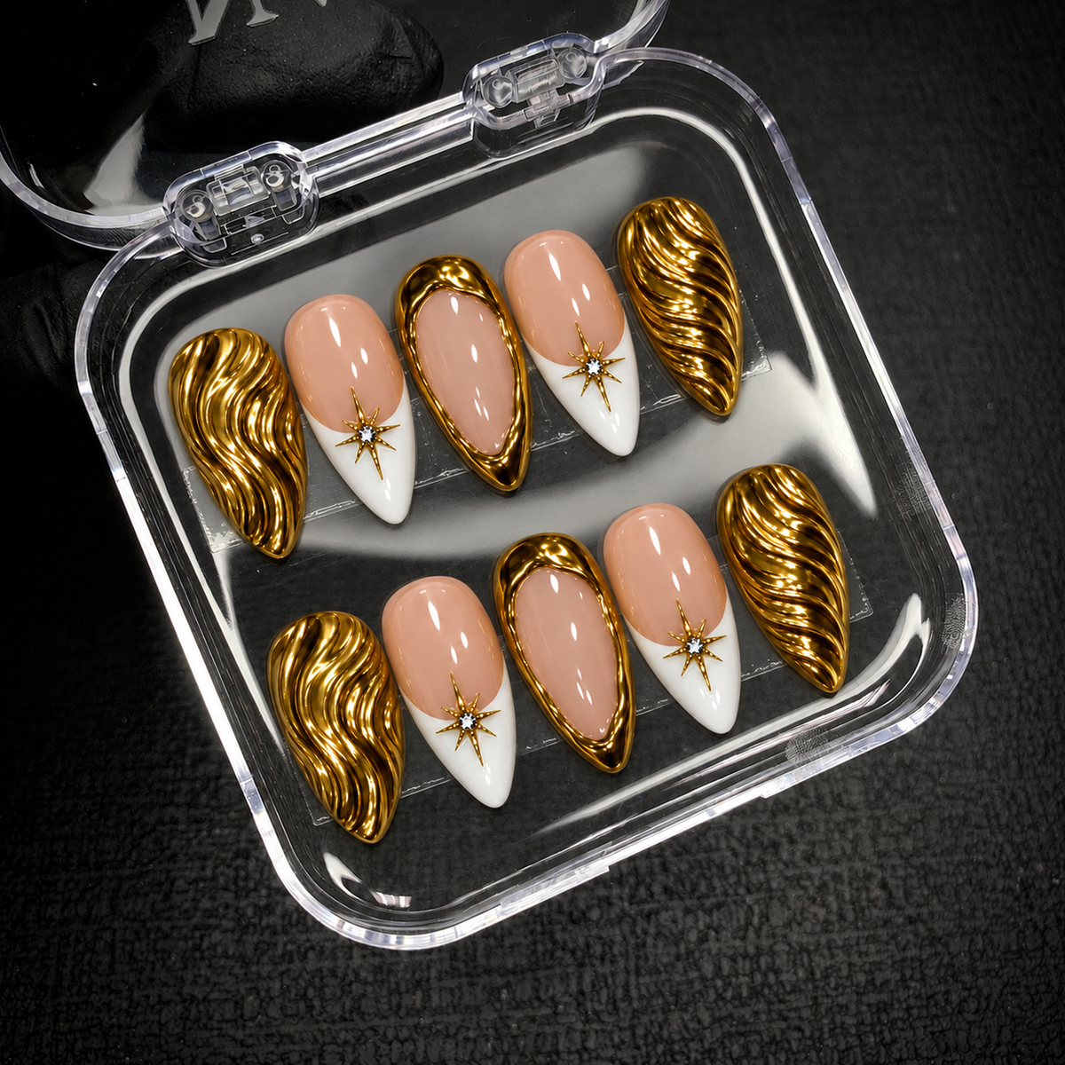 Open clear nail case on dark textured fabric holding ten peach, white, and gold nails with wave patterns, gold borders, and starburst accents.