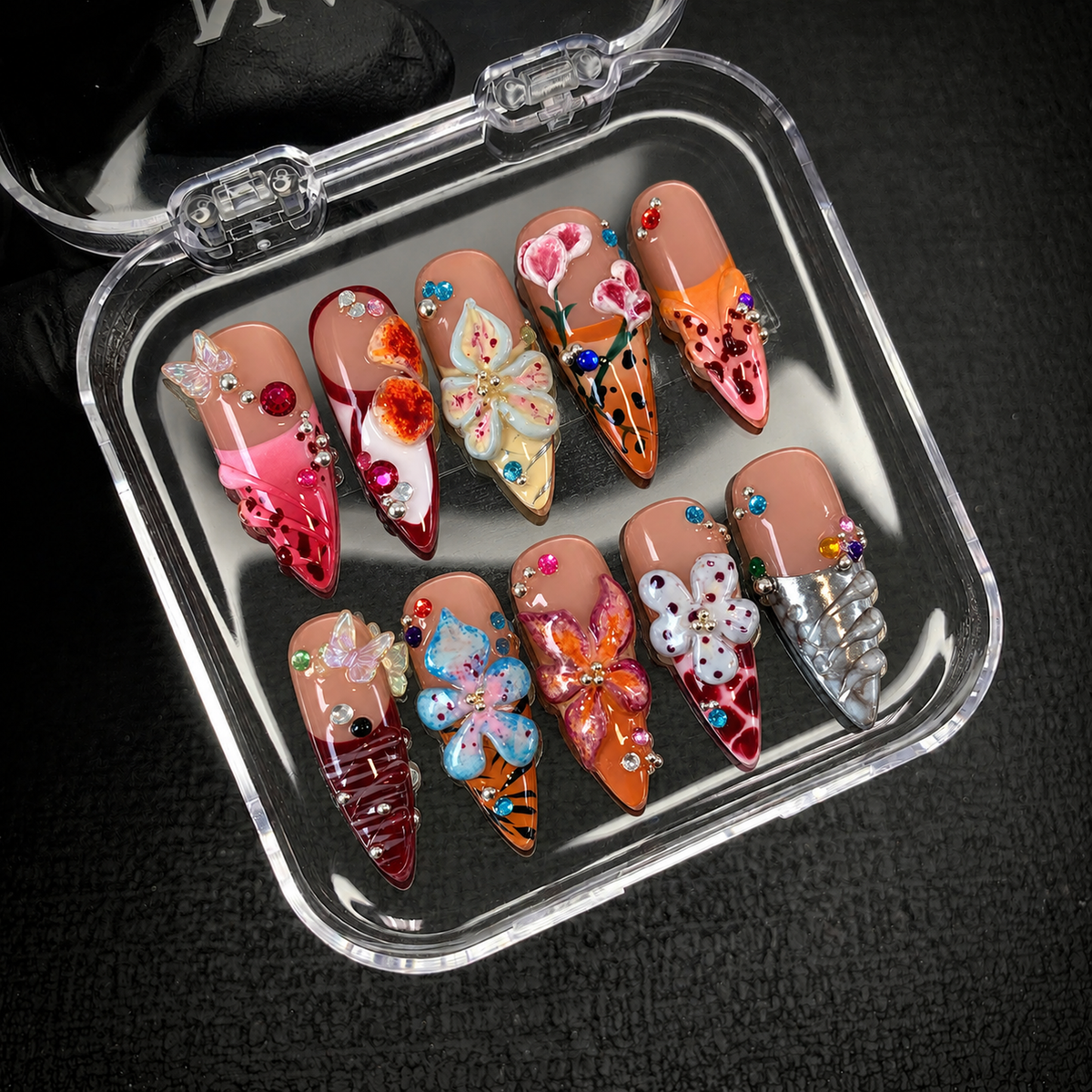 Open clear case displaying ten coordinated floral and butterfly accent nails against a dark textured background.