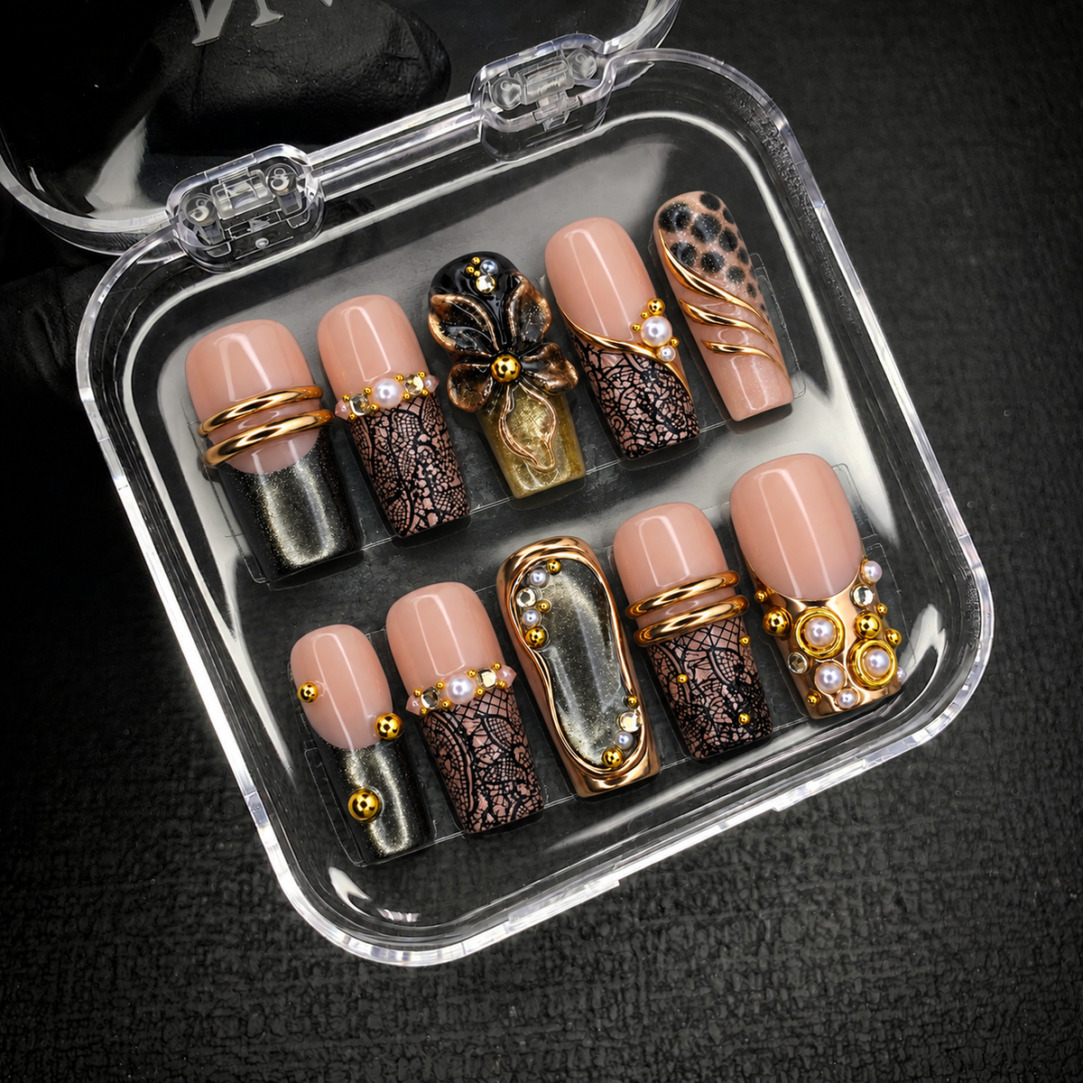 Open clear case holding ten decorated nails on a dark textured background, with floral, lace-like, shimmer, and gold-tone details visible.