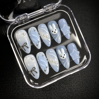 Open clear case displaying ten coordinated pale blue press-on nails with bunny, star, bow, and pearl details.