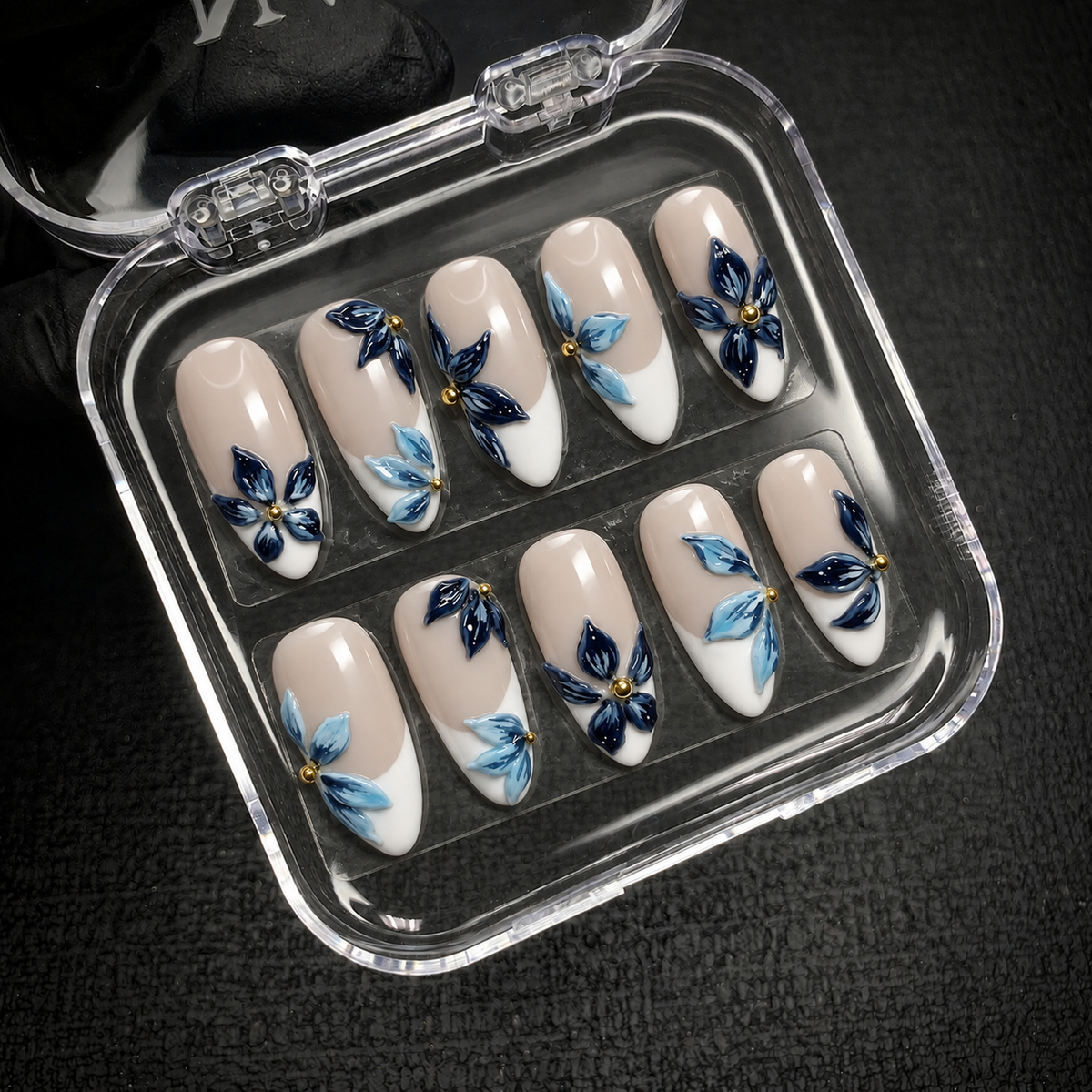 Open clear nail case on a dark textured surface holding ten nude and white French nails decorated with blue flowers and gold-tone accents.