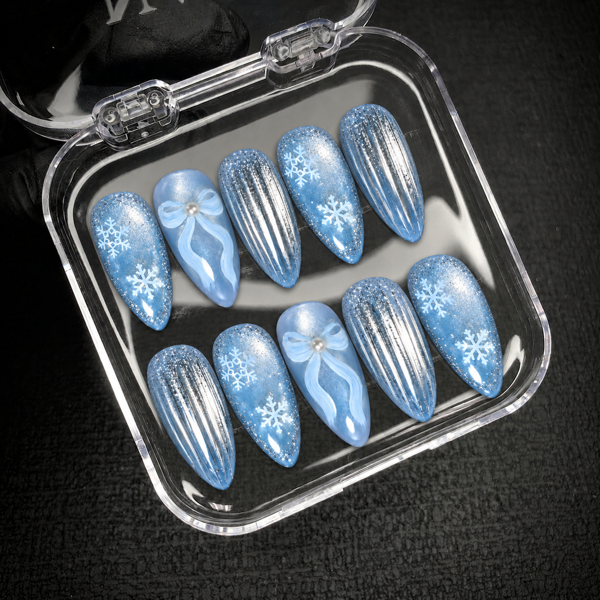 Open clear nail case displaying ten icy blue nails with snowflakes, pale blue bows, and silver stripes against a dark background.