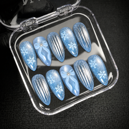Open clear nail case displaying ten icy blue nails with snowflakes, pale blue bows, and silver stripes against a dark background.