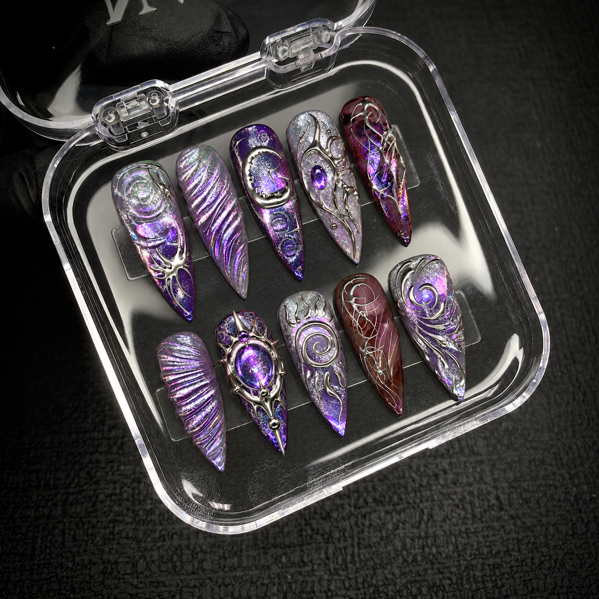 Open clear case holding ten purple and silver decorated nails in two rows on a dark textured surface.