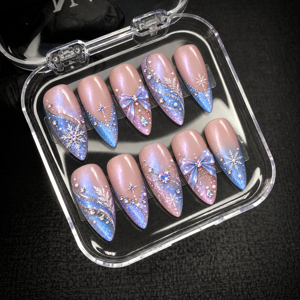 Open clear nail case on a dark textured surface holding ten coordinated pink, blue, and lavender nails with bows, snowflakes, and gem accents.