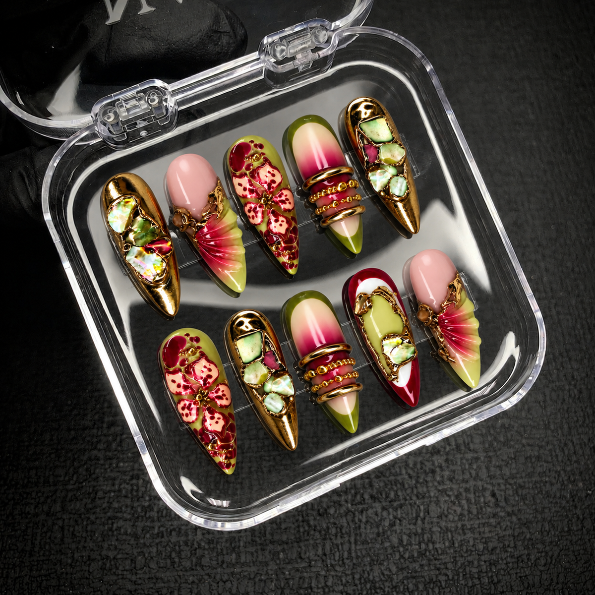Open clear case holding ten coordinated floral and mosaic nails with pink, olive, berry, and gold details against a dark textured background.