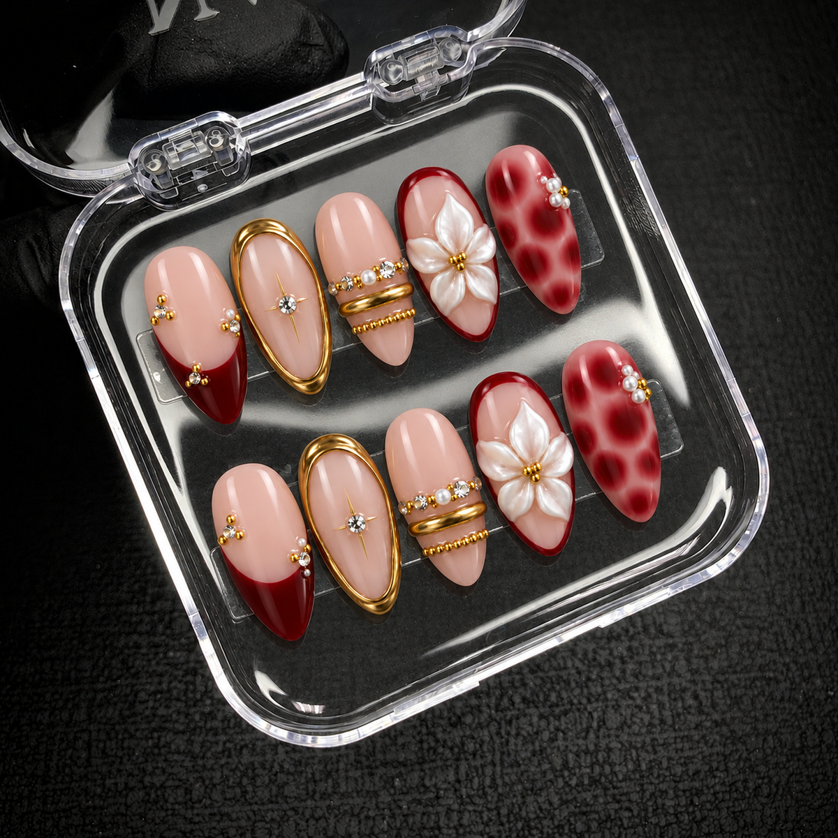 Open clear case holding ten coordinated blush pink and deep red nails with raised white flowers and gold-tone accents on a dark textured surface.