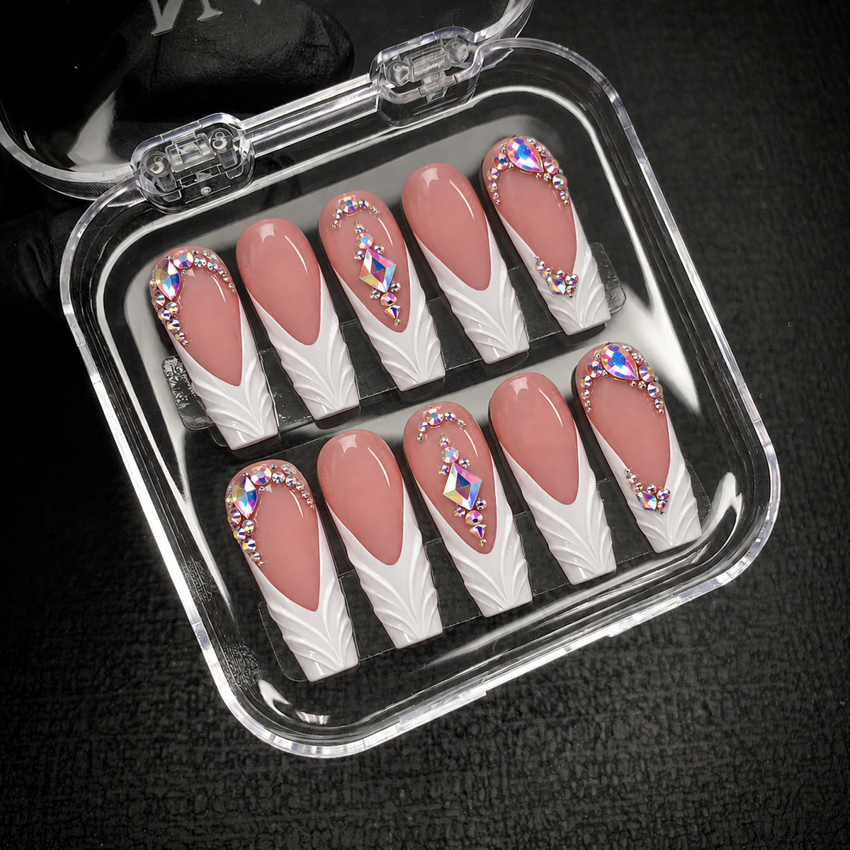 Open clear case holding ten coordinated dusty rose and white nails with raised tip detailing and iridescent gems on a dark textured surface.
