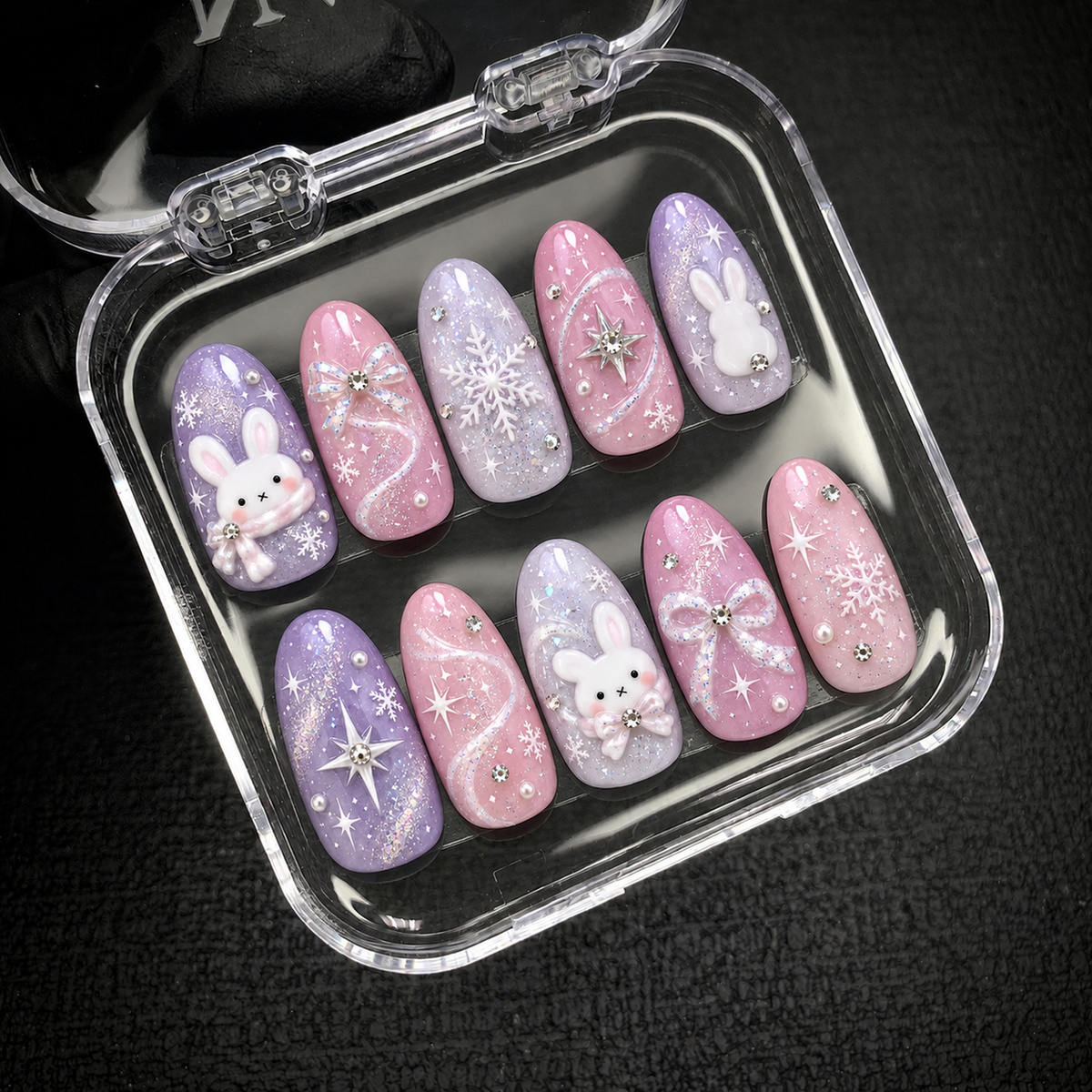 Open clear nail case on dark textured fabric holding ten pastel winter nails with white bunnies, snowflakes, bows, and glitter details.