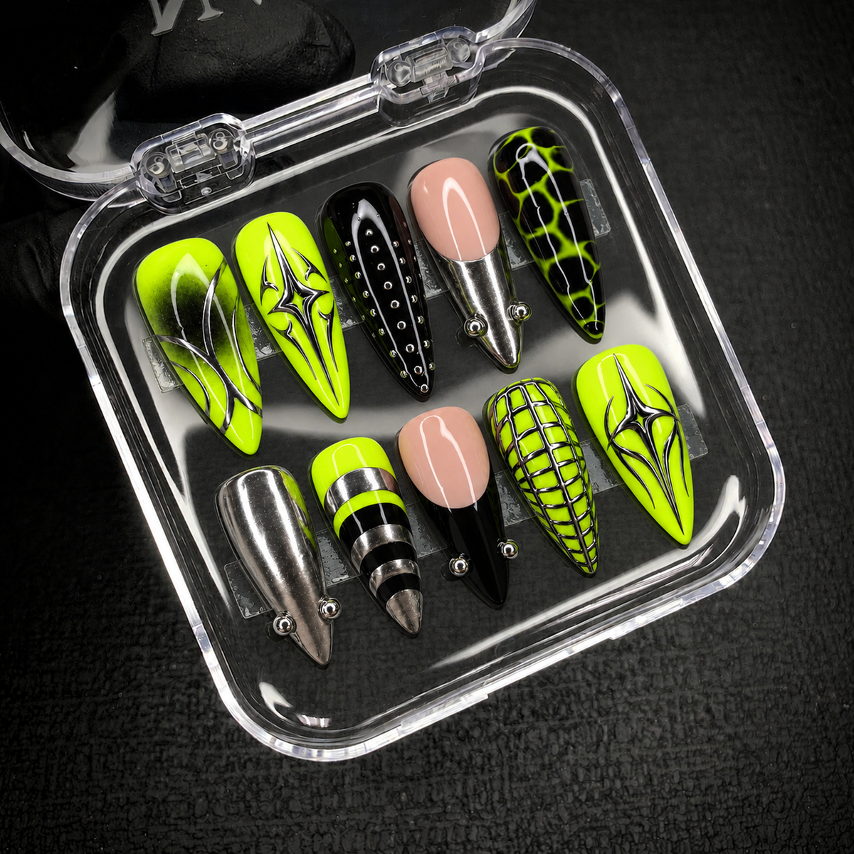 Open clear nail case on a dark textured background holding ten neon lime, black, nude pink, and silver nails arranged in two rows.
