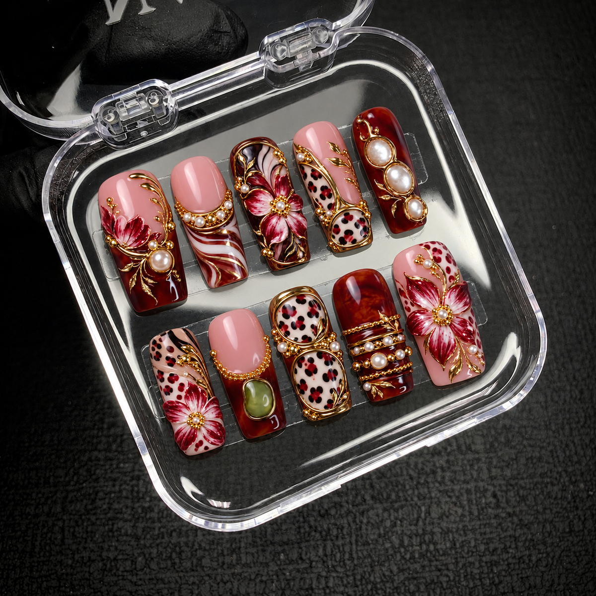 Open clear nail case on a dark textured surface, displaying ten burgundy and blush nails with floral artwork, leopard spots, and gold-tone decorative details.
