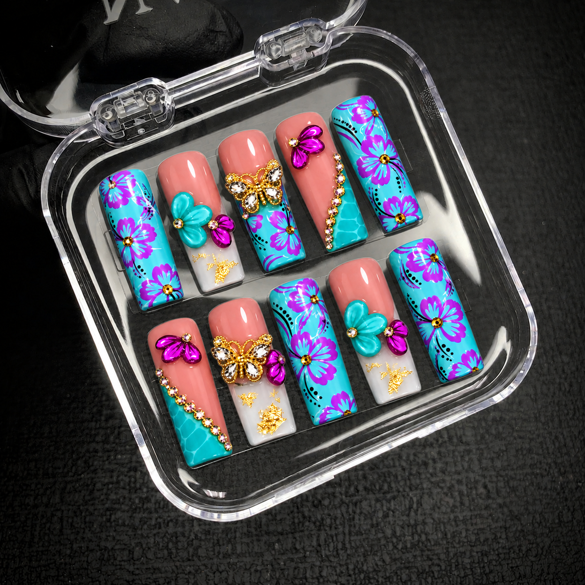 Open clear case on a dark textured background holding ten turquoise floral and coral pink nails with raised petals, jeweled butterflies, and gold-tone details.