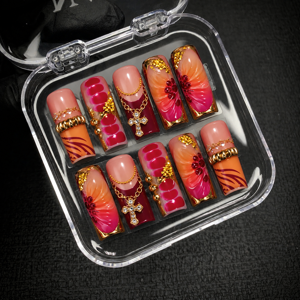 Open clear case holding ten decorated nails on dark textured fabric, showing floral accents, gold-tone borders, chains, and cross charms.