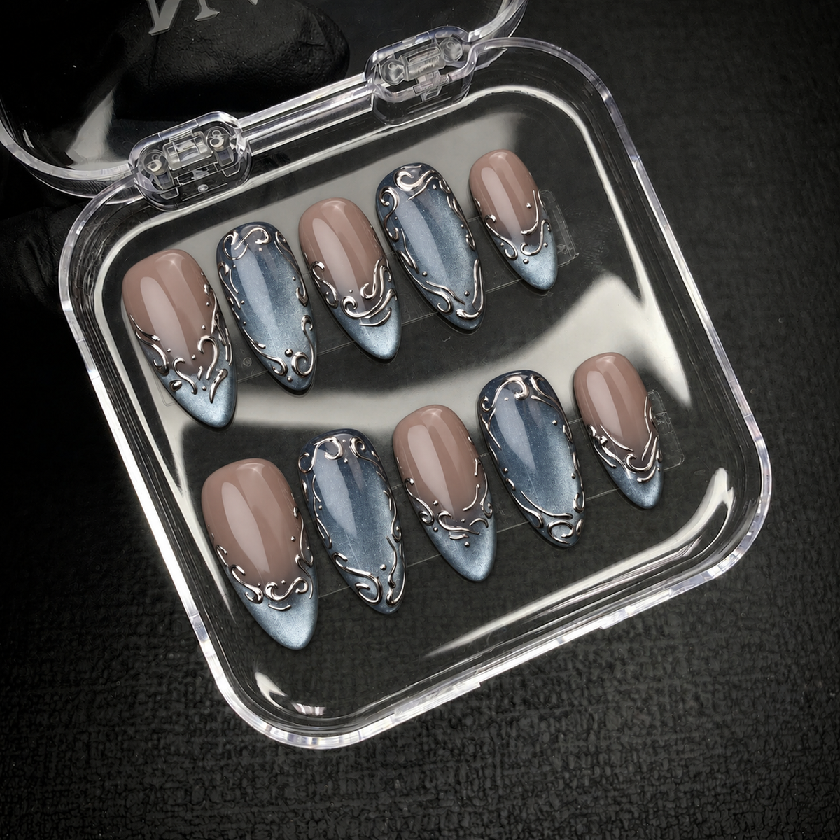 Open clear nail case displaying ten coordinated greige and blue-gray nails with silver-toned scrollwork against a dark background.