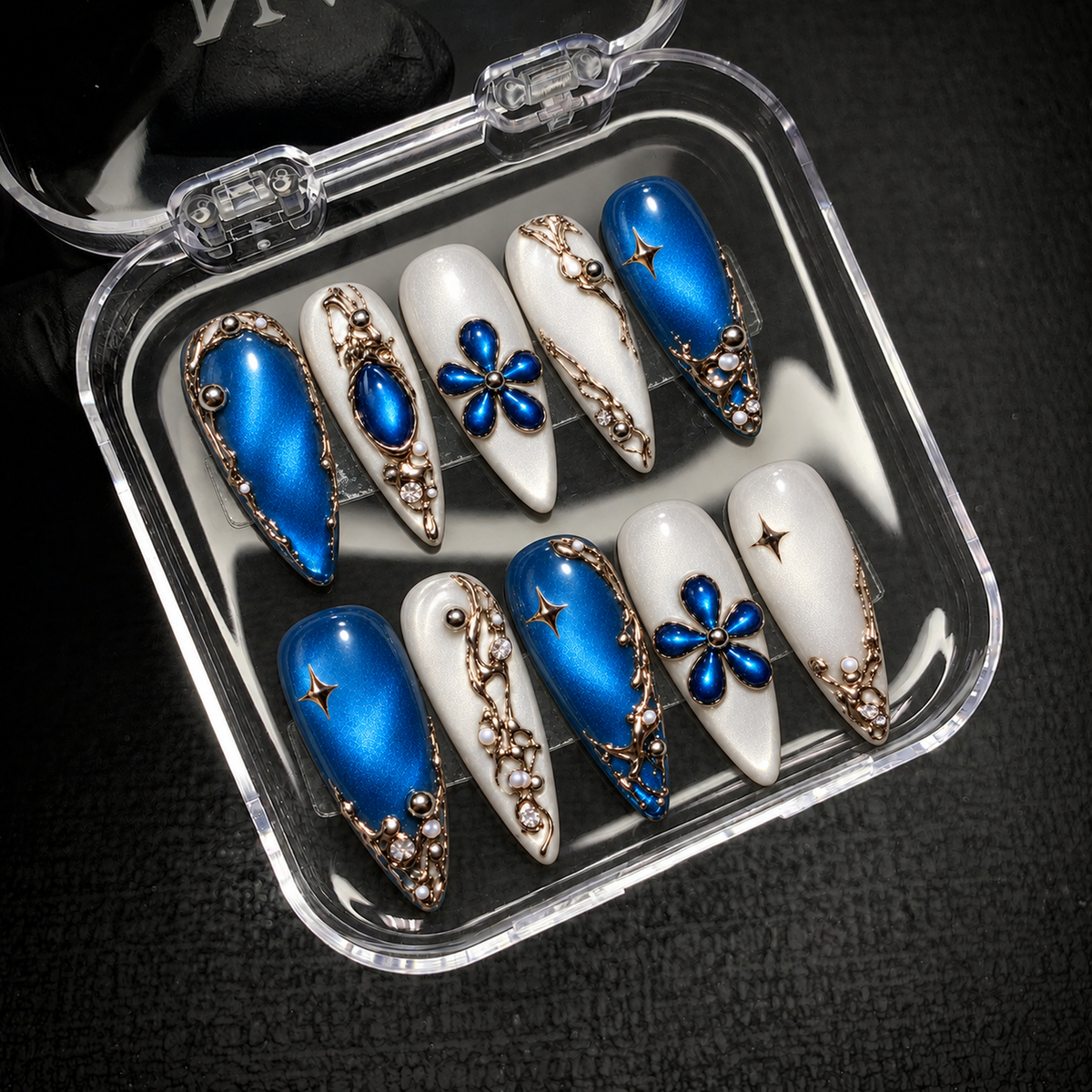 Open clear case displaying ten coordinated blue and white nails with floral, star, and metallic-toned accents.