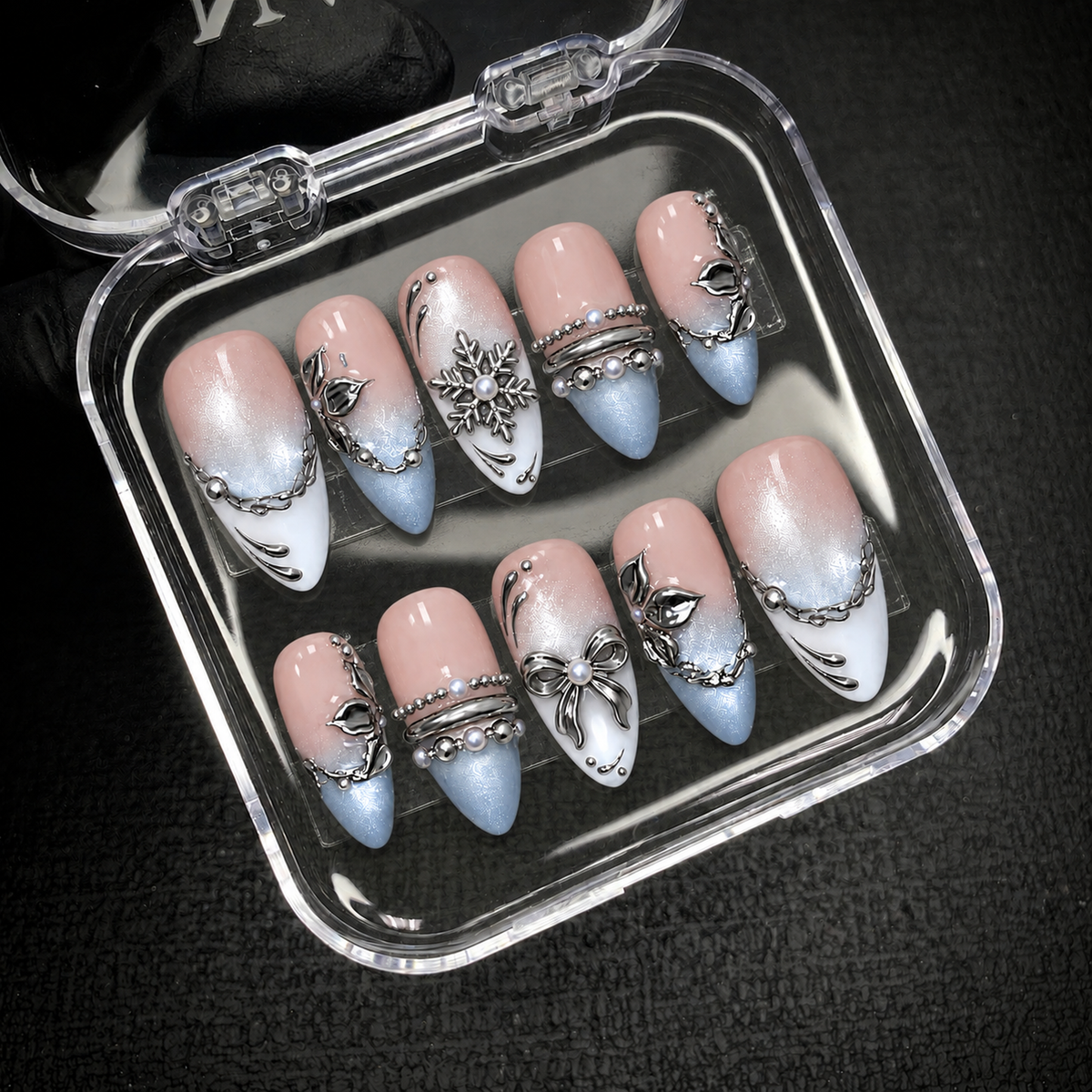 Ten blush pink, icy blue, and white nails with silver-tone details displayed inside an open clear case against a dark background.