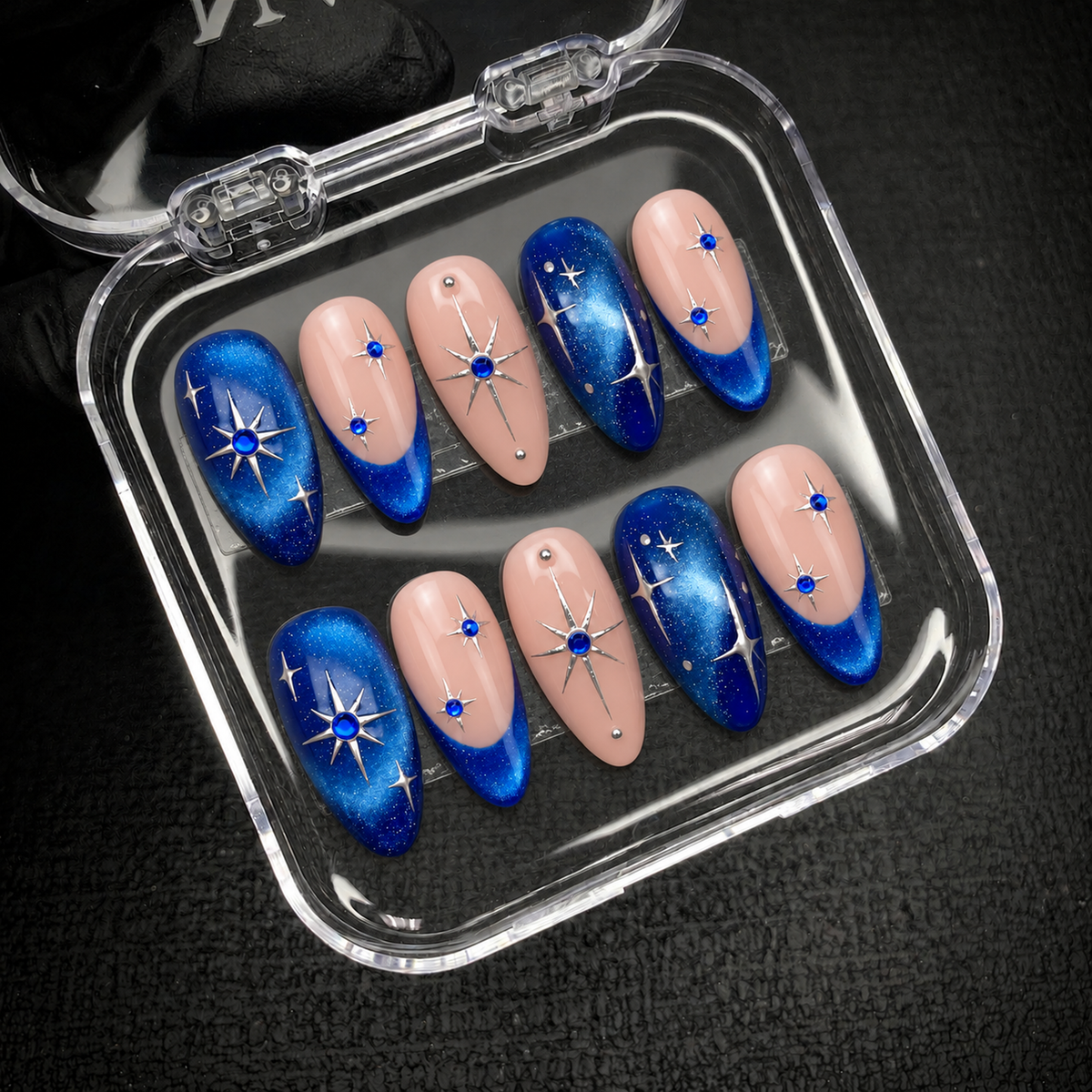Open clear nail case on dark textured fabric displaying ten blush pink and blue shimmer nails with silver stars and blue gem-like accents.