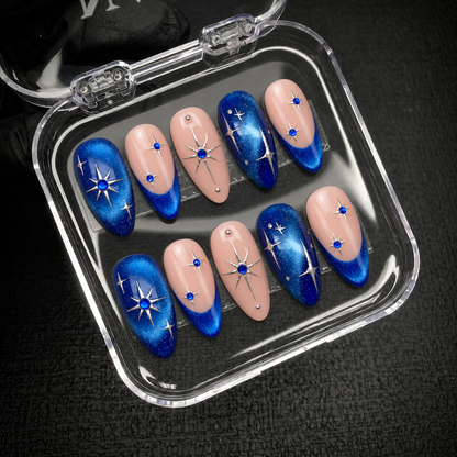 Open clear nail case on dark textured fabric displaying ten blush pink and blue shimmer nails with silver stars and blue gem-like accents.
