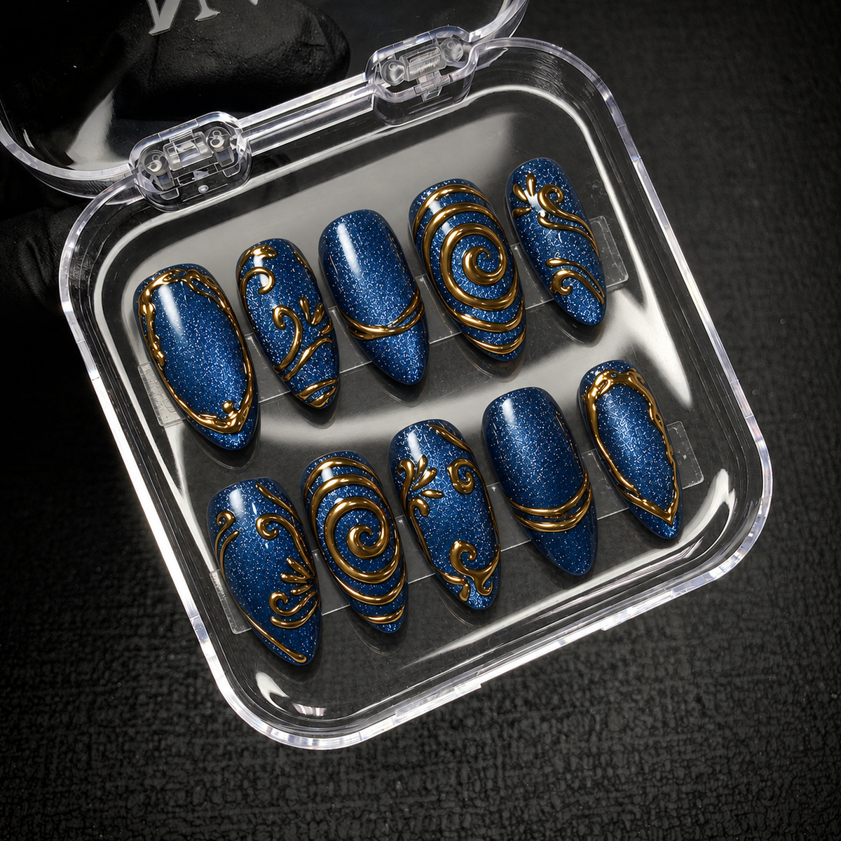 Open clear case displaying ten deep blue shimmer nails with gold ornamental details against a dark background.