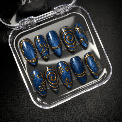 Open clear case displaying ten deep blue shimmer nails with gold ornamental details against a dark background.