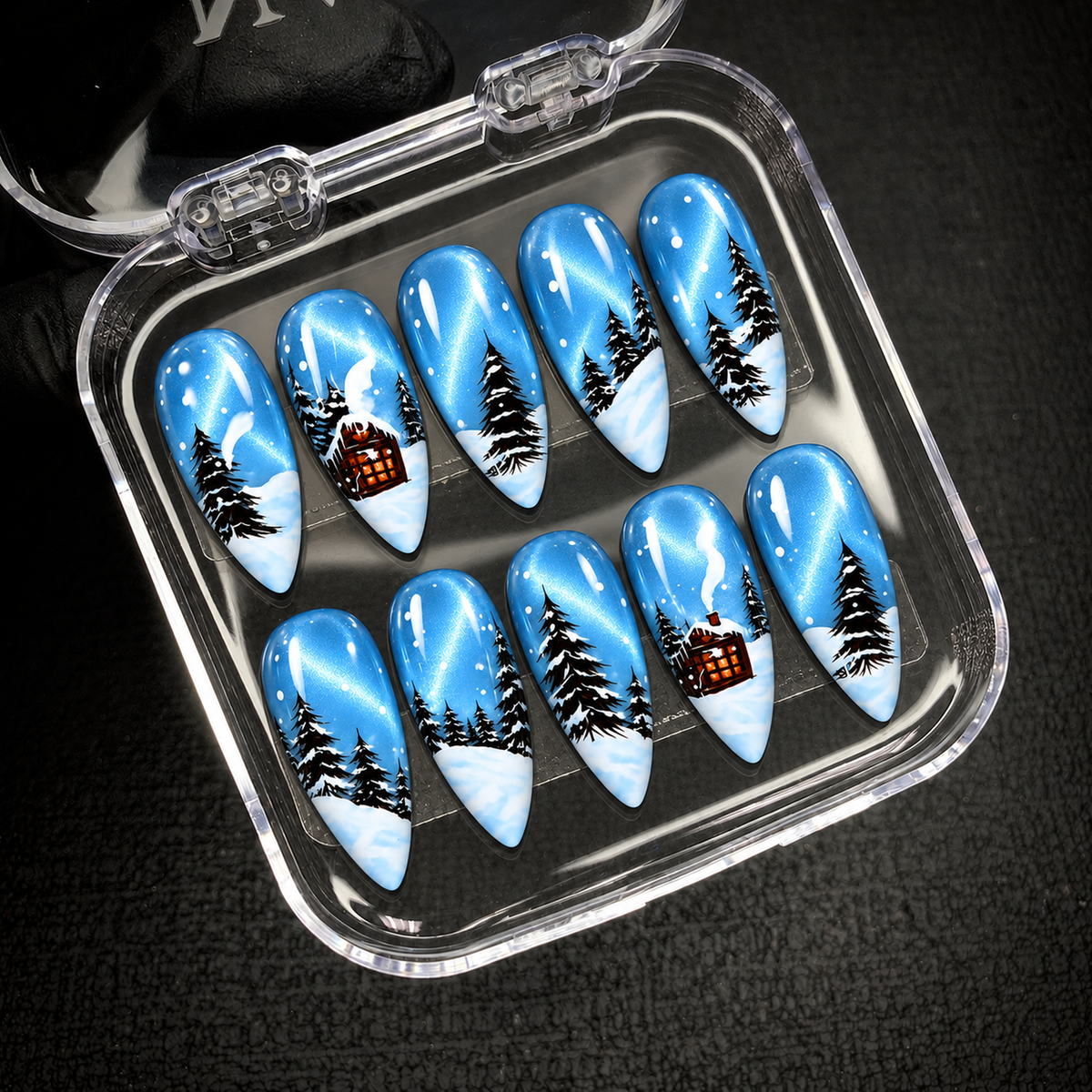Open clear nail case on a dark textured surface, holding ten blue nails decorated with snowy trees and warm orange cabin windows.