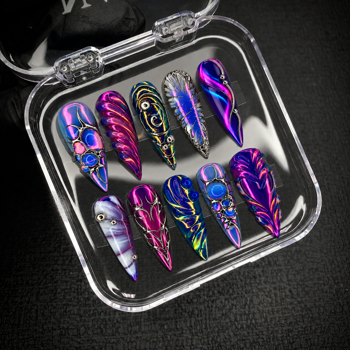 Open clear nail case on a dark textured background displaying ten colorful pointed nails with silver-tone linework and sculptural-looking accents.