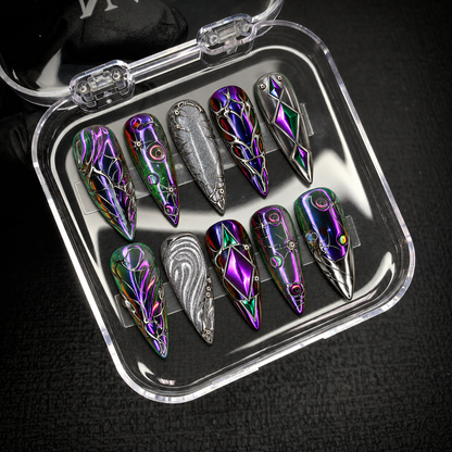 Open clear case on a dark textured surface holding ten coordinated nails with purple and green reflections, silver scrollwork, and geometric diamond details.