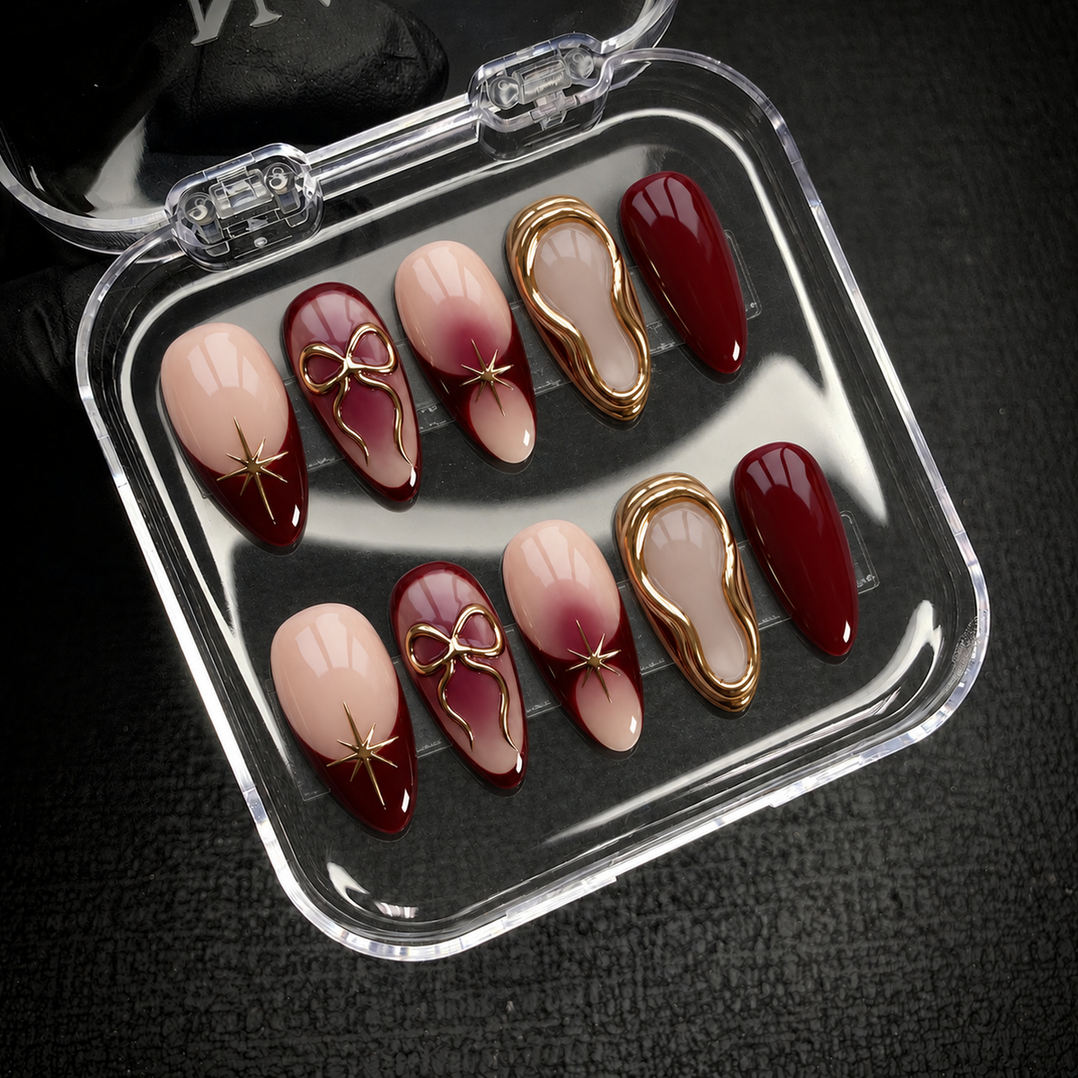 Open clear nail case on dark textured fabric holding ten coordinated burgundy and nude nails with gold decorative accents.