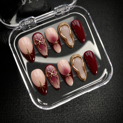Open clear nail case on dark textured fabric holding ten coordinated burgundy and nude nails with gold decorative accents.