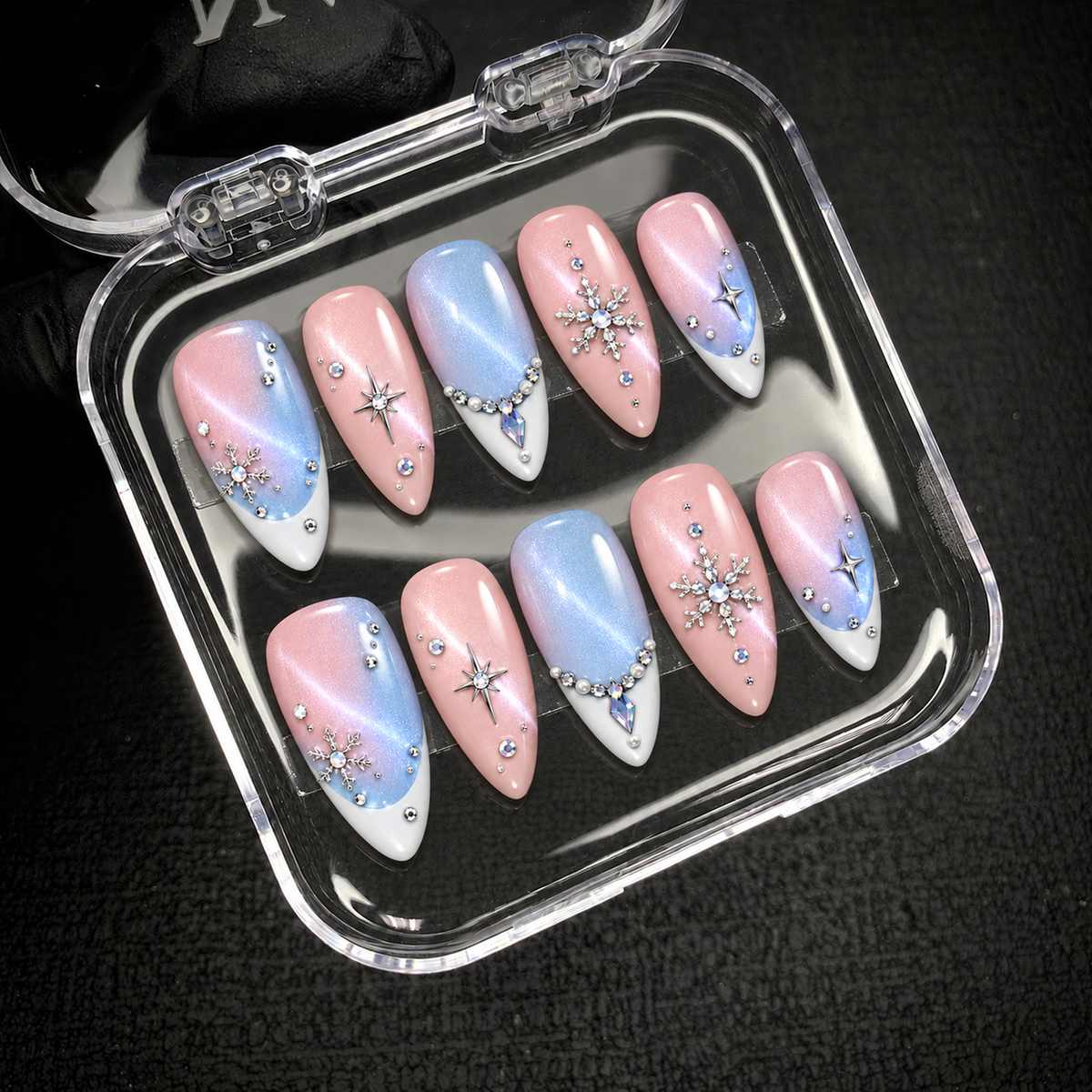 Open clear case on a dark textured surface holding ten pink and icy blue nails with shimmering gradients, white tips, snowflakes, and starbursts.