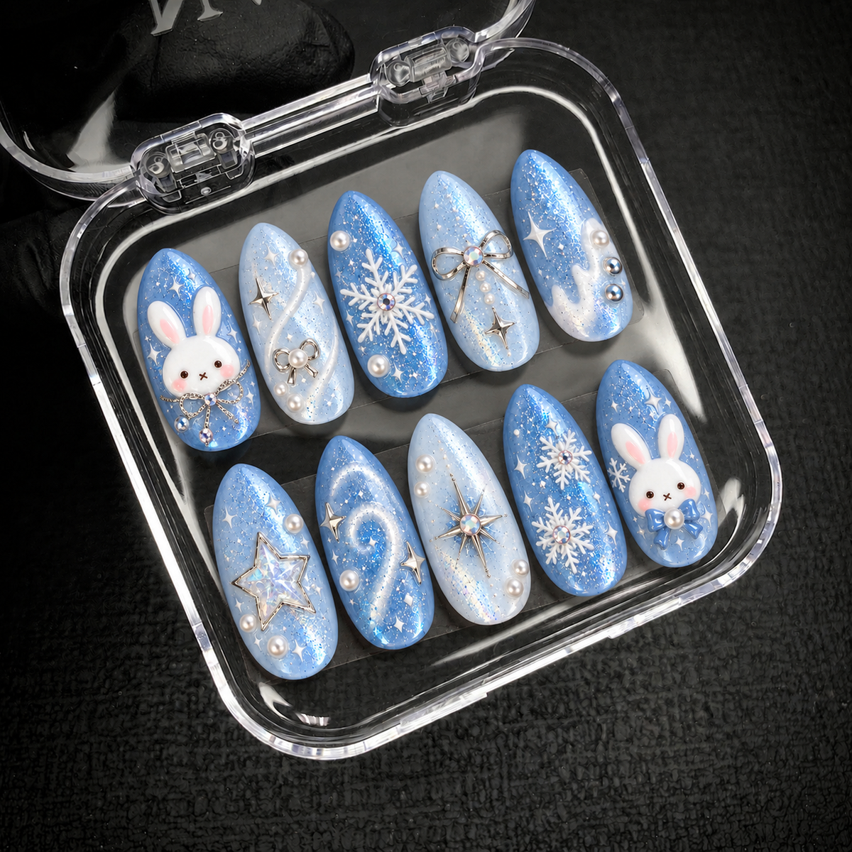 Open clear nail case on a dark textured surface holding ten blue nails decorated with bunny faces, white snowflakes, bows, and star accents.