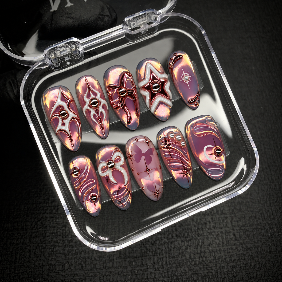 Open clear nail case on dark textured fabric displaying ten mauve iridescent nails with metallic and pale outlined motifs.
