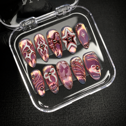 Open clear nail case on dark textured fabric displaying ten mauve iridescent nails with metallic and pale outlined motifs.