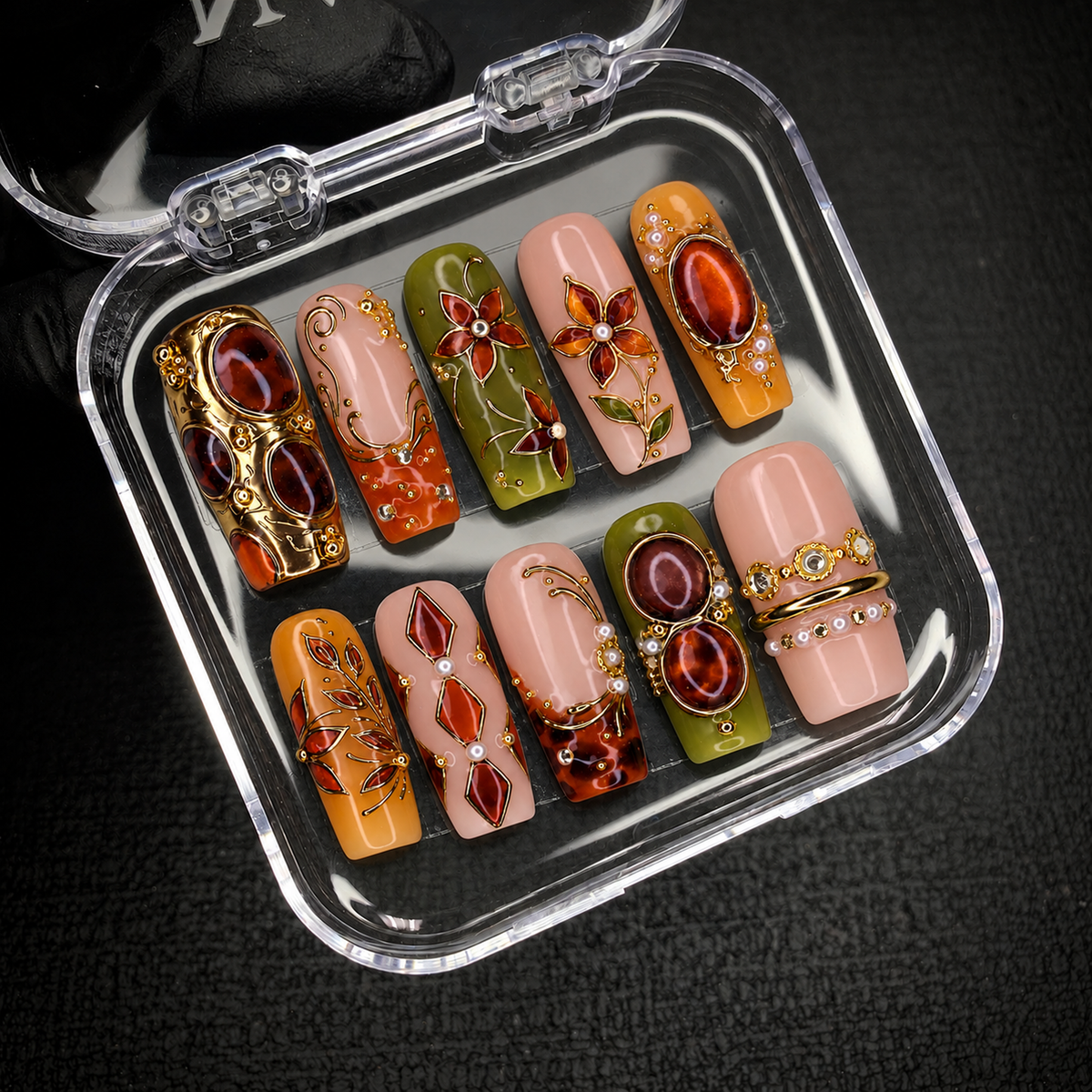 Open clear nail case on a dark textured surface holding ten coordinated nails with floral motifs, amber-toned accents, and gold-tone ornamentation.