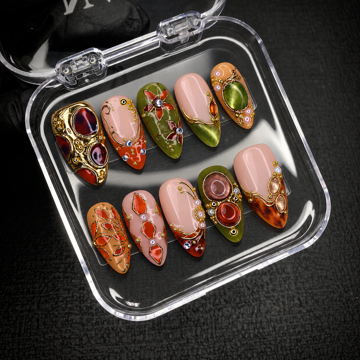 Open clear case holding ten coordinated botanical nails with gold-tone detailing on a dark textured background.