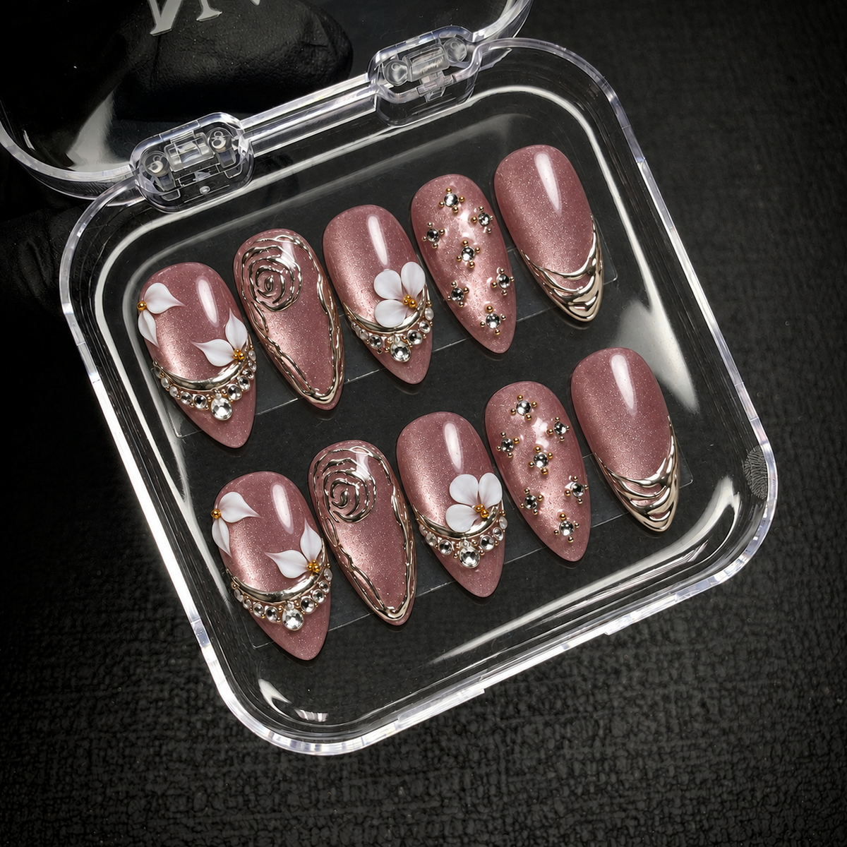 Open clear nail case on a dark textured surface holding ten mauve shimmer nails with white flowers, silver-toned accents, and clear gems.