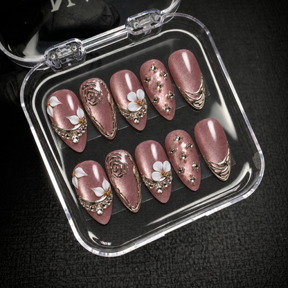 Open clear nail case on a dark textured surface holding ten mauve shimmer nails with white flowers, silver-toned accents, and clear gems.