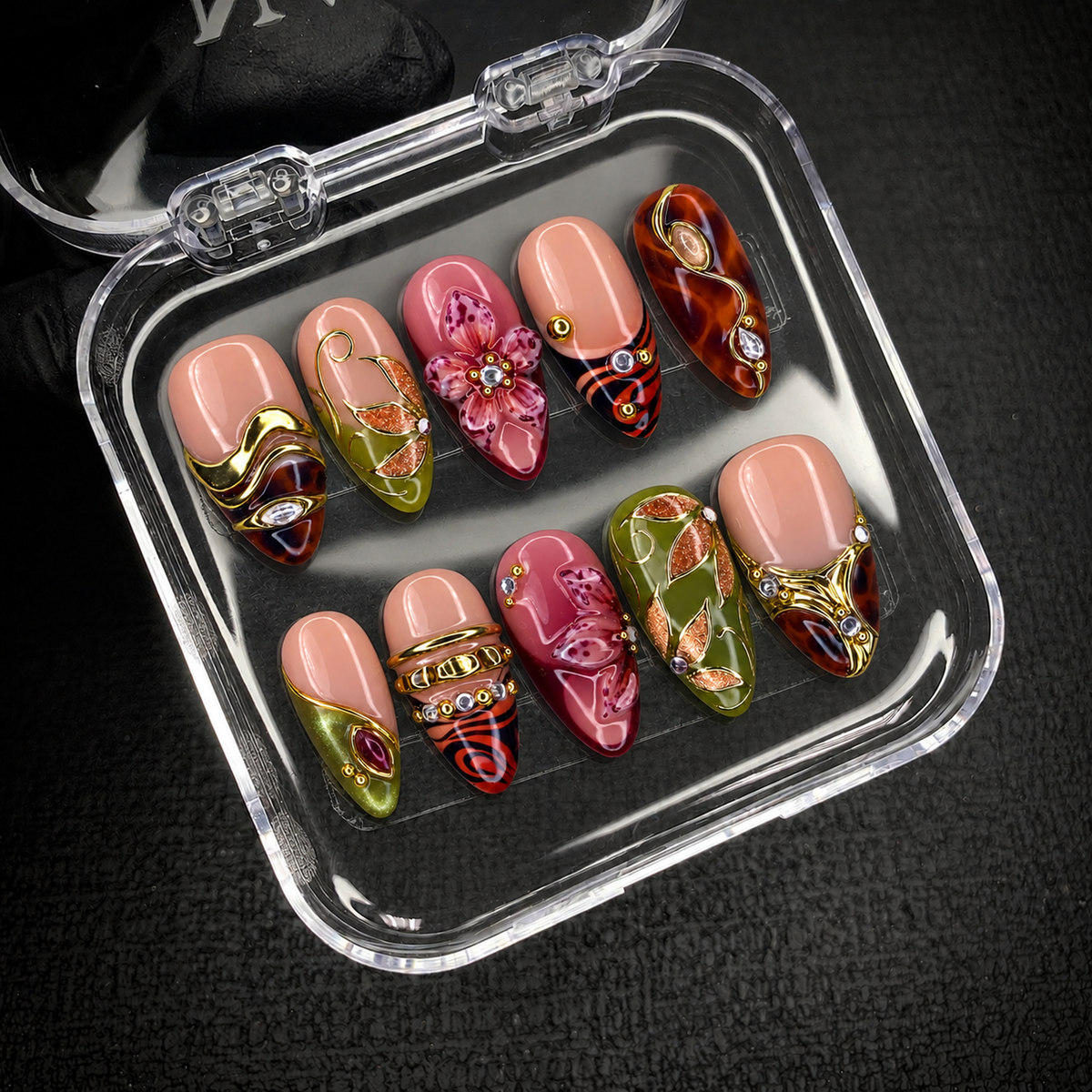 Open clear case holding ten decorated nails with pink flowers, olive leaf motifs, and gold-toned details against a dark textured background.