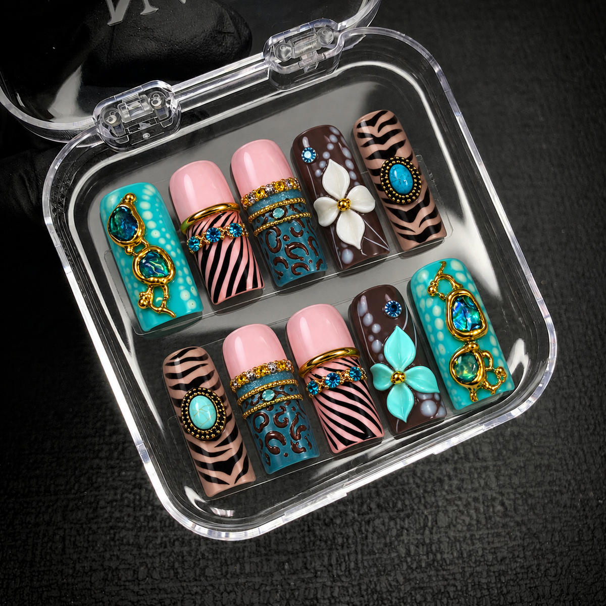 Open clear case holding ten coordinated nails with turquoise embellishments, striped and spotted patterns, and white and aqua flowers on dark fabric.