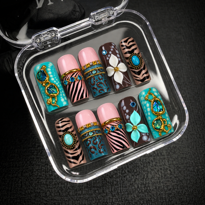 Open clear case holding ten coordinated nails with turquoise embellishments, striped and spotted patterns, and white and aqua flowers on dark fabric.