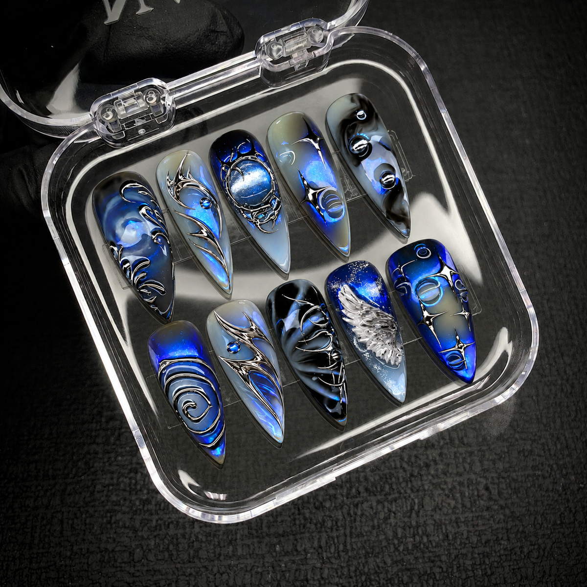 Open clear case on a dark textured surface holding ten coordinated nails with cobalt shimmer and silver celestial accents.