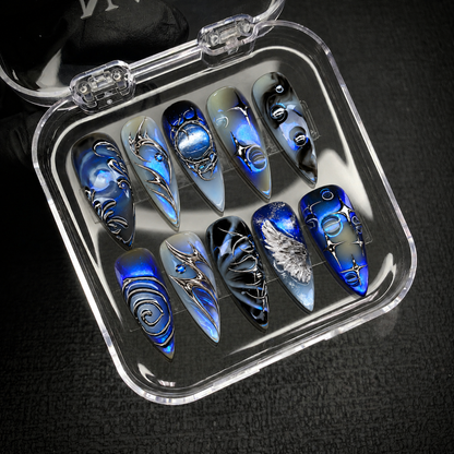 Open clear case on a dark textured surface holding ten coordinated nails with cobalt shimmer and silver celestial accents.