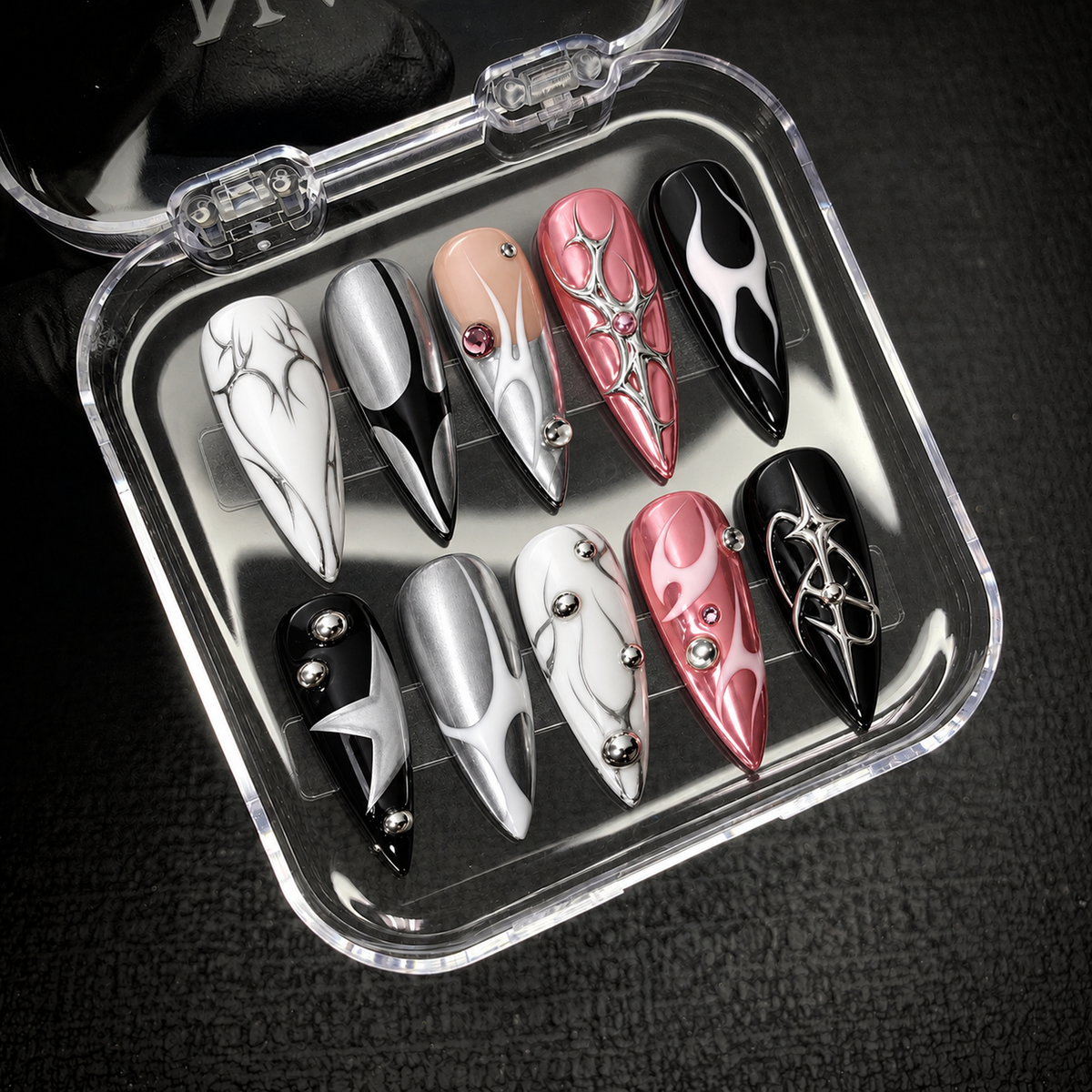 Open clear case holding ten coordinated nails with pink and silver effects, black-and-white graphics, and raised embellishments on a dark textured surface.