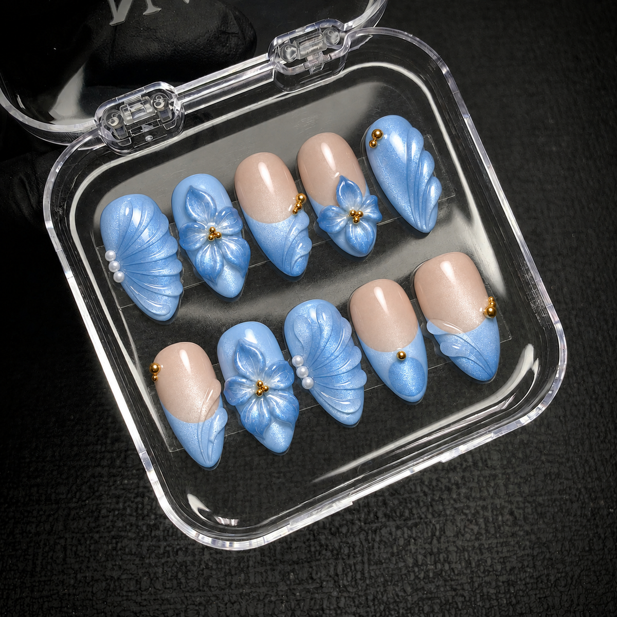 Ten blue and nude floral press-on nails arranged inside an open clear case against a dark background.
