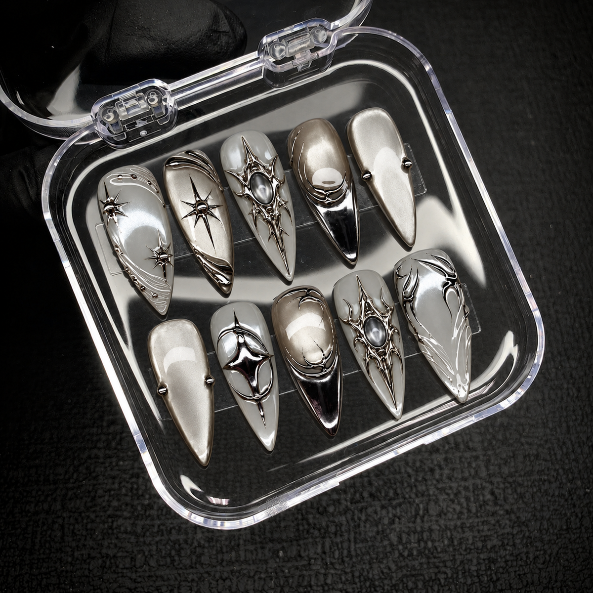 Open clear case holding ten coordinated silver and gray pointed nails with starbursts and chrome ornamentation on black textured fabric.