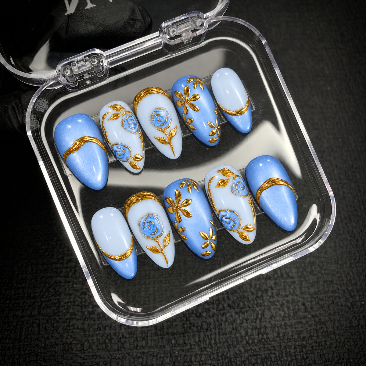Open clear case holding ten coordinated blue floral nails with gold rose outlines, leaves, and curved bands on a dark textured surface.