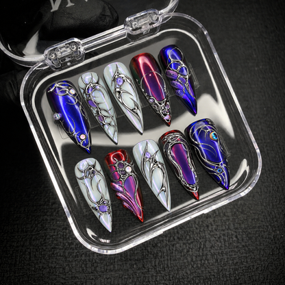 Open clear case on dark textured fabric displaying ten coordinated pointed nails with jewel-tone and pearly bases and silver-tone linework.
