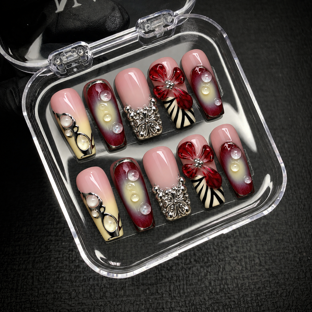 Open clear case holding ten coordinated pink, burgundy, and cream-yellow nails with red flowers and silver-tone ornament on a dark textured surface.