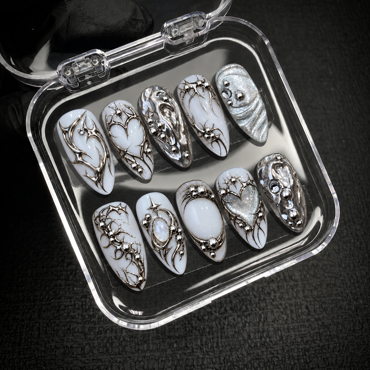 Open clear nail case on a dark textured surface displaying ten coordinated white and silver nails with chrome swirls, hearts, and starbursts.