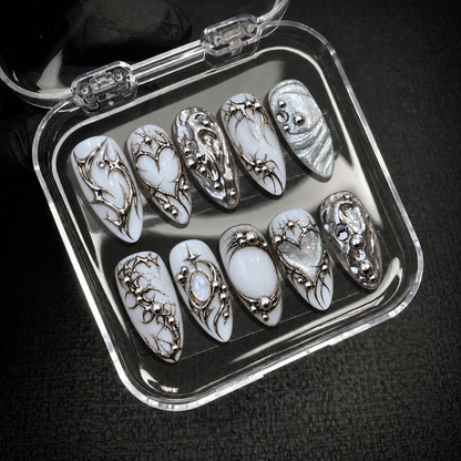 Open clear nail case on a dark textured surface displaying ten coordinated white and silver nails with chrome swirls, hearts, and starbursts.